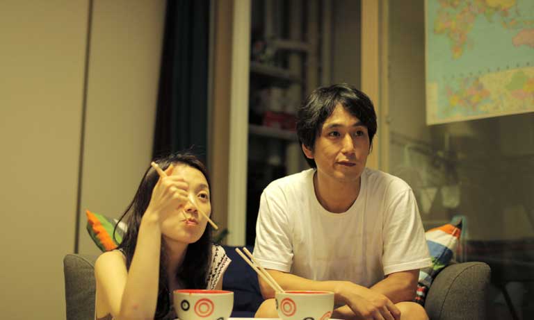 A slightly bored-looking couple watch TV while eating noodles