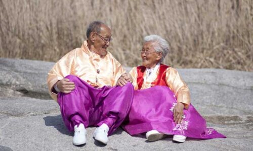A lughing elderly couple in matchin hanbok sit on a rock