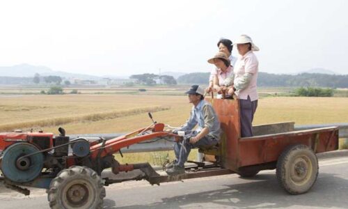 Three women ride across farmland in a trailer attached to a small tractor