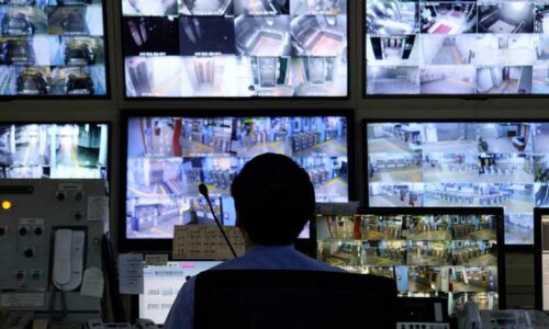 A silhouette of a man watching a myriad of CCTV monitors