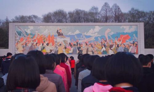 Girls stand in front of a mural of Kim Il Sung