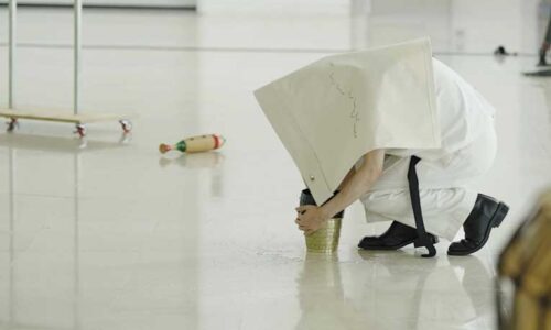 Meta-Hierarchical Exercise - an artist-performer crouches under a white cloth embroidered with an abstract musical score