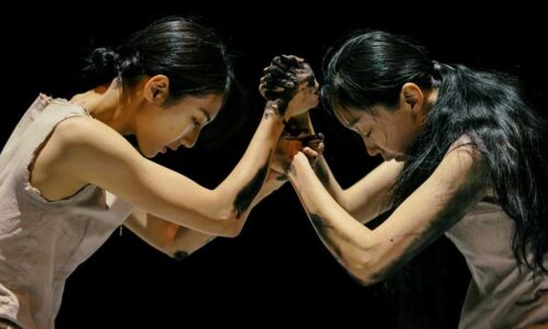 Two dancers stained with ash lean against each other