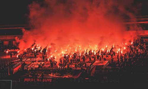 Night-time shot of football supporters carrying red flaming torches in the stands of a stadium