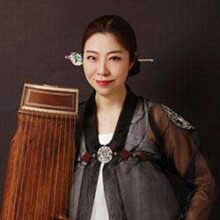 Ji Eun Jung has her hair up and stands next to her kayagum which is a Korean traditional harp.